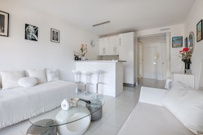 A modern, white-themed living area with a sofa, a glass coffee table, and a kitchen in the background.