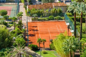 Una cancha de tenis rodeada de abundante vegetación y un edificio con techo de tejas.