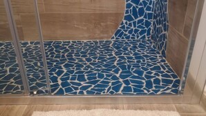 Une salle de bain avec un mur de douche recouvert de mosaïques bleues, un tapis blanc et moelleux orné de motifs de voiliers, et un plancher en bois.