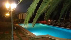 Une piscine illuminée la nuit, avec des palmiers et des chaises longues.