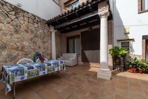 Un patio con una mesa cubierta con mantel, una silla blanca, plantas en macetas y un muro de piedra.