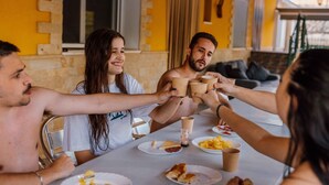 Un grupo de personas brindando alrededor de una mesa con comida y bebidas, en una habitación con paredes amarillas y una ventana.