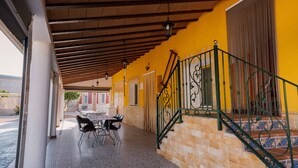 Un espacio al aire libre techado con una mesa y sillas, una escalera con barandillas decorativas y una pared amarilla.