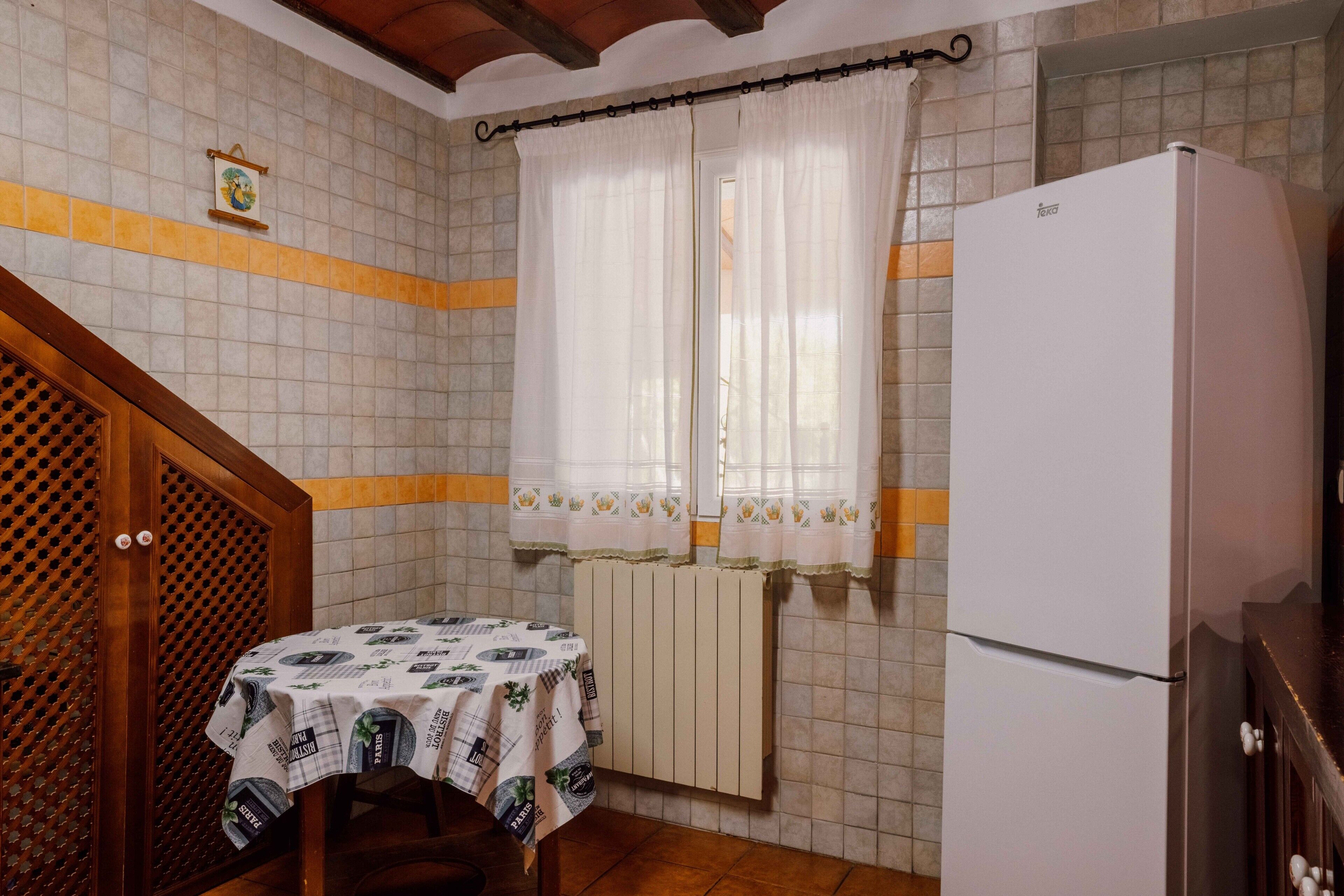 Una cocina con refrigerador, una mesa con mantel, una ventana con cortinas y una pared con azulejos.