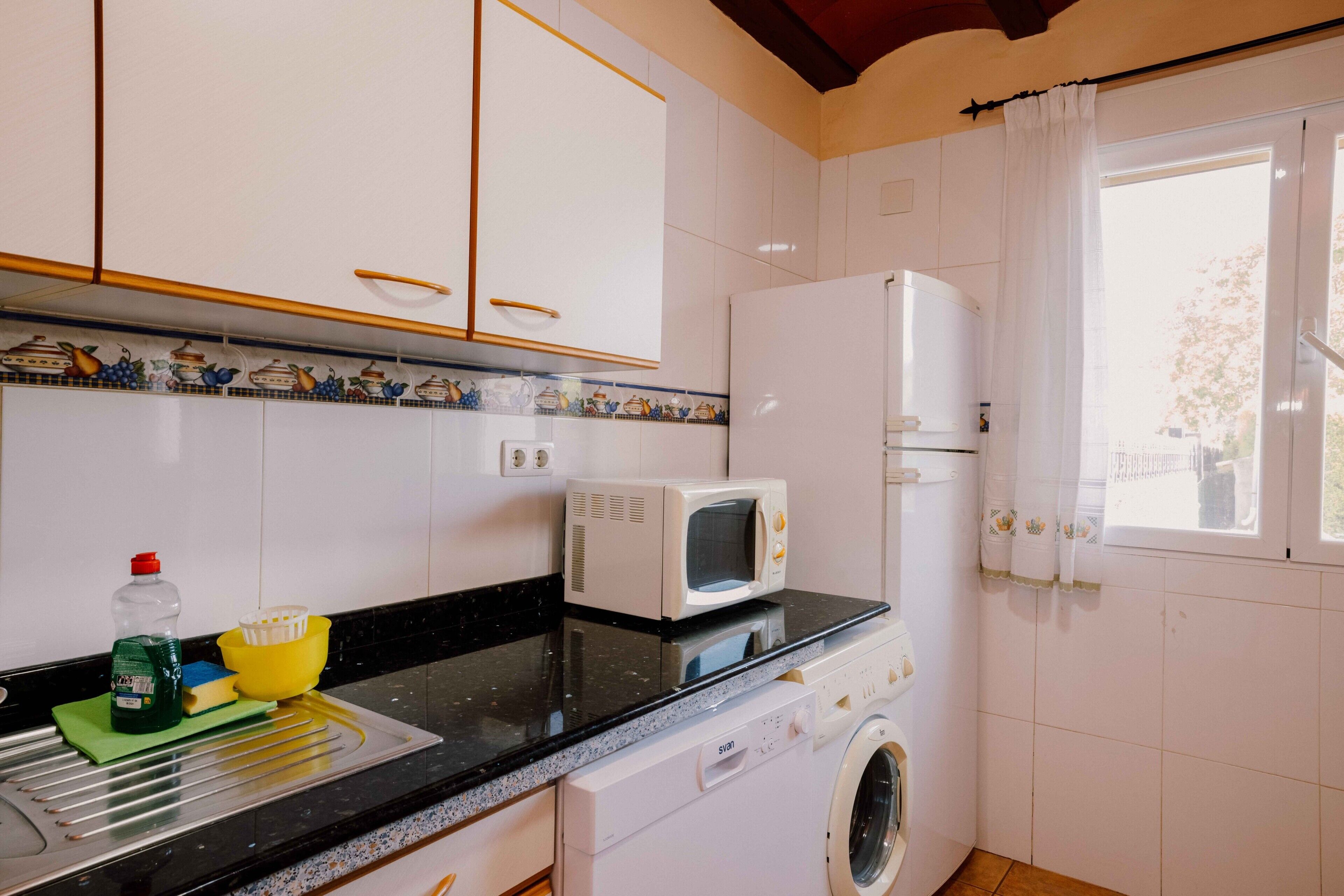 Una cocina equipada con lavadora, microondas y una ventana con cortinas.