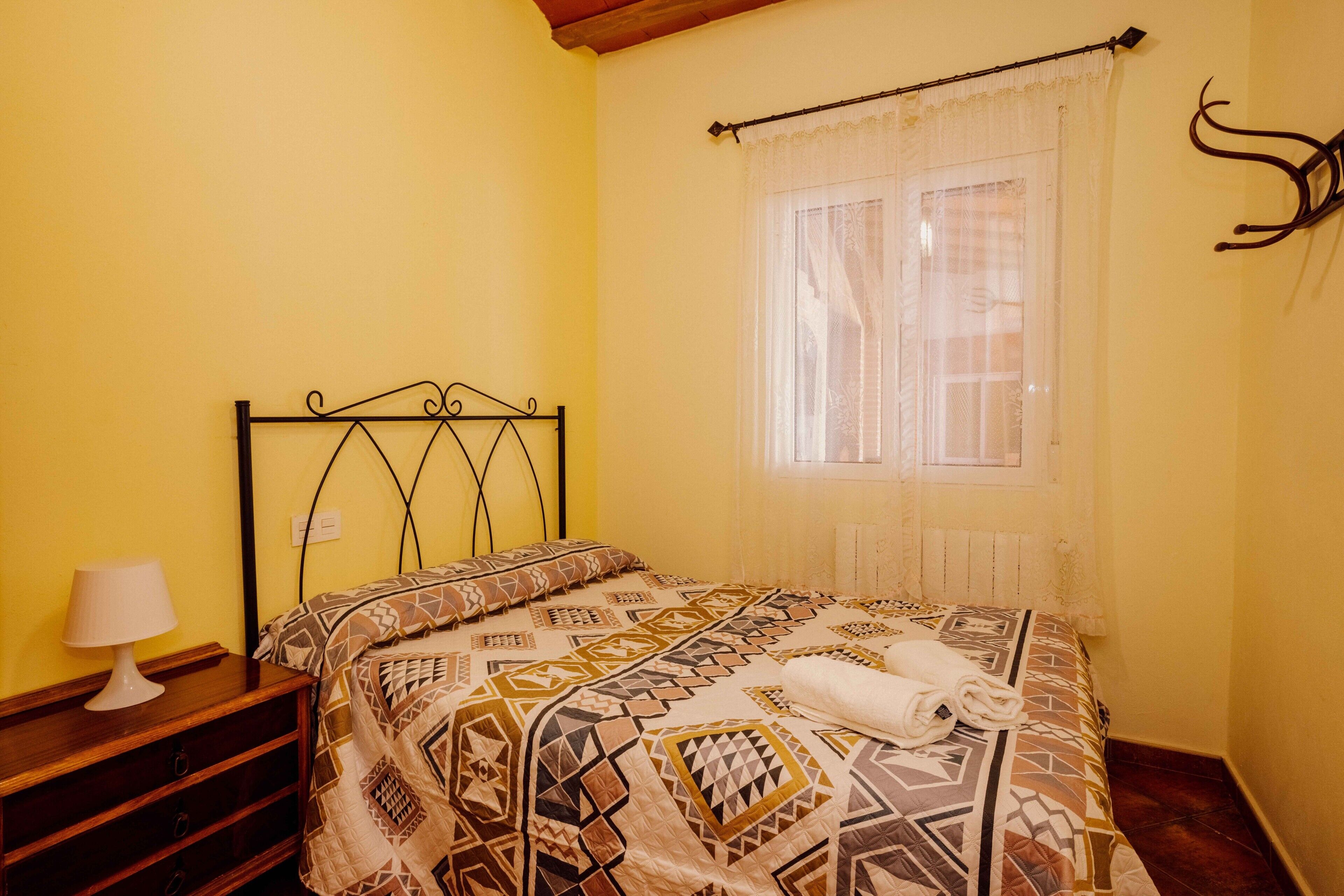 A bedroom with a bed, a nightstand, a lamp, and a window with a curtain.