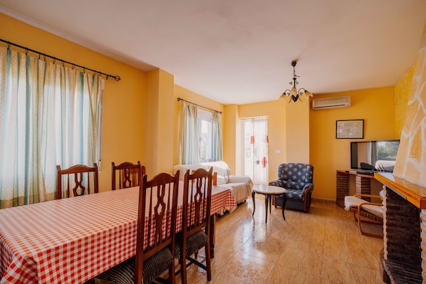 Una habitación con una mesa a cuadros, sillas de madera, un sofá, una mesita redonda, un televisor y una chimenea.