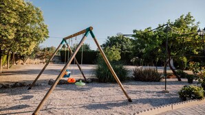 A swing set in a park with gravel ground, surrounded by trees and plants.
