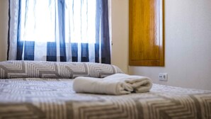 A neatly made bed with patterned bedding, a window with sheer curtains, and a wooden cabinet on the wall.