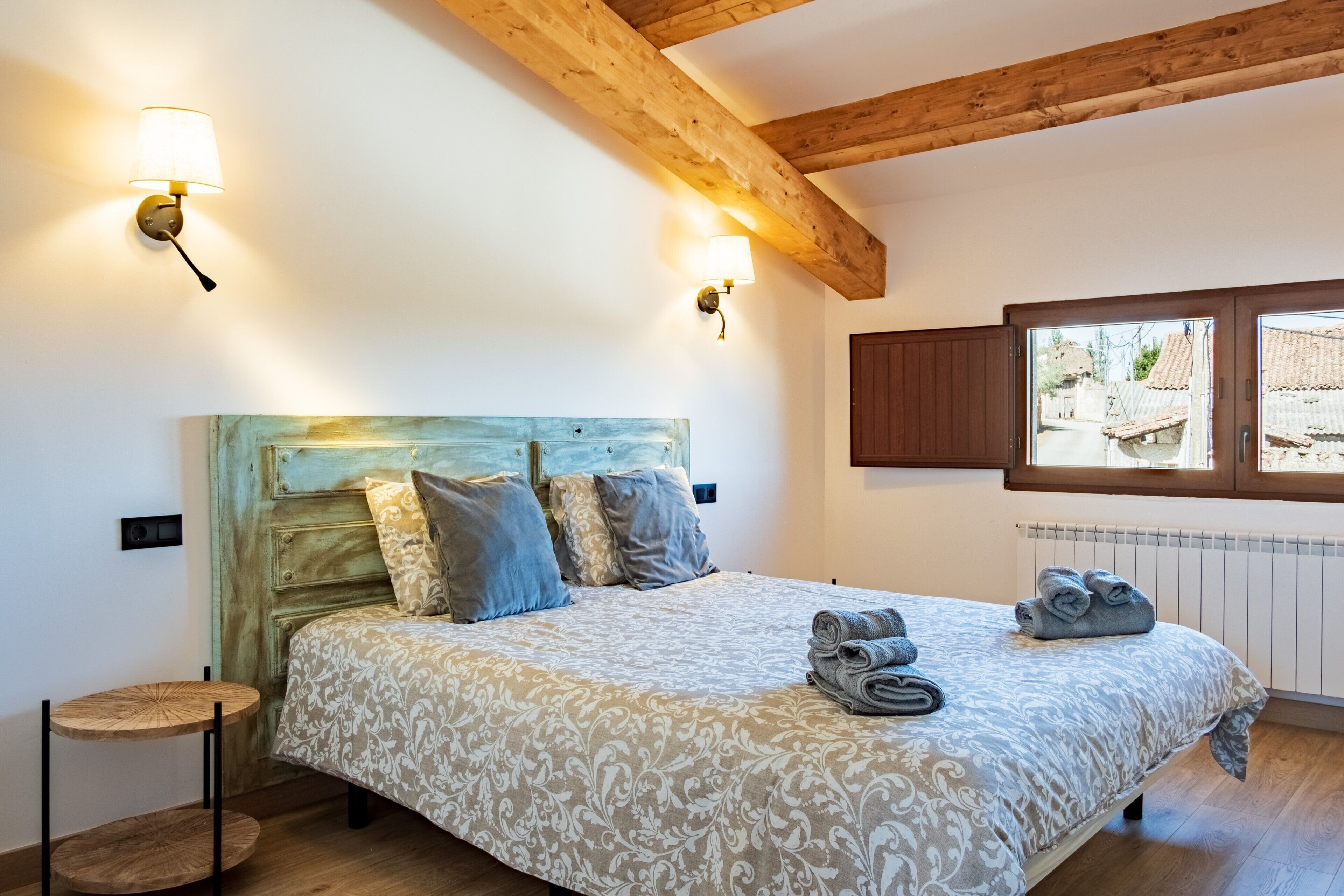 Een slaapkamer met een bed, twee wandlampen, een houten hoofdbord, een klein rond tafeltje en een raam met uitzicht op gebouwen.