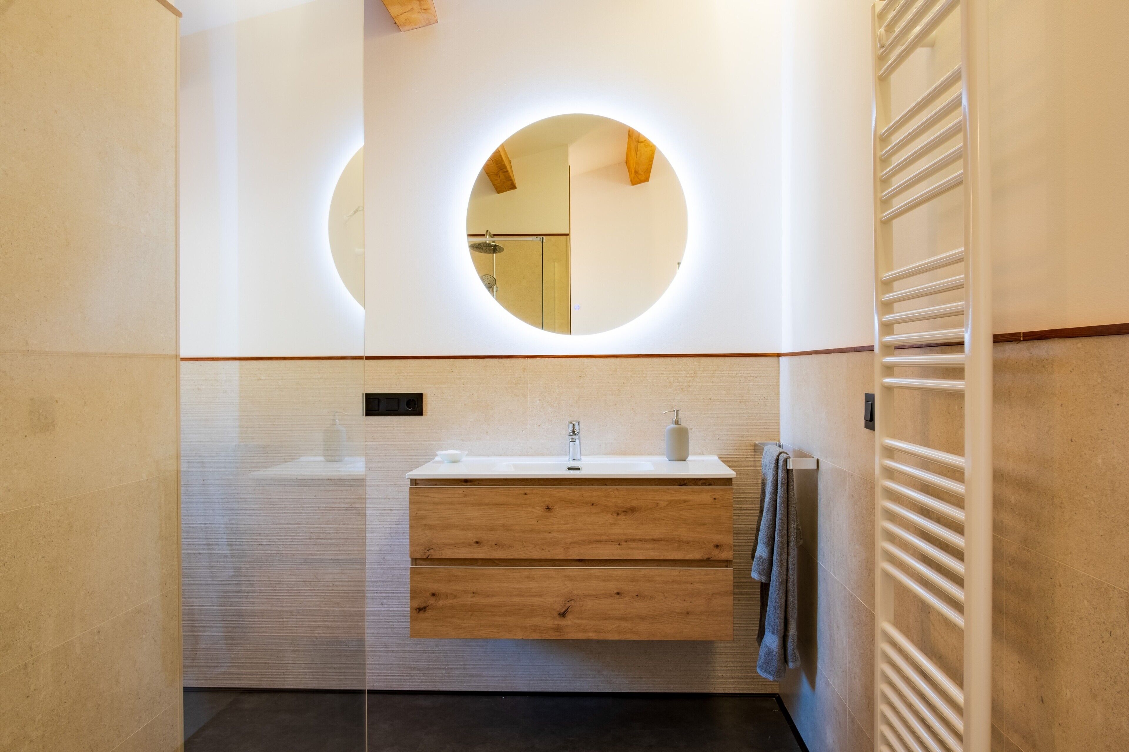 Une salle de bain moderne avec un grand miroir rond, un meuble-lavabo en bois et un porte-serviettes.