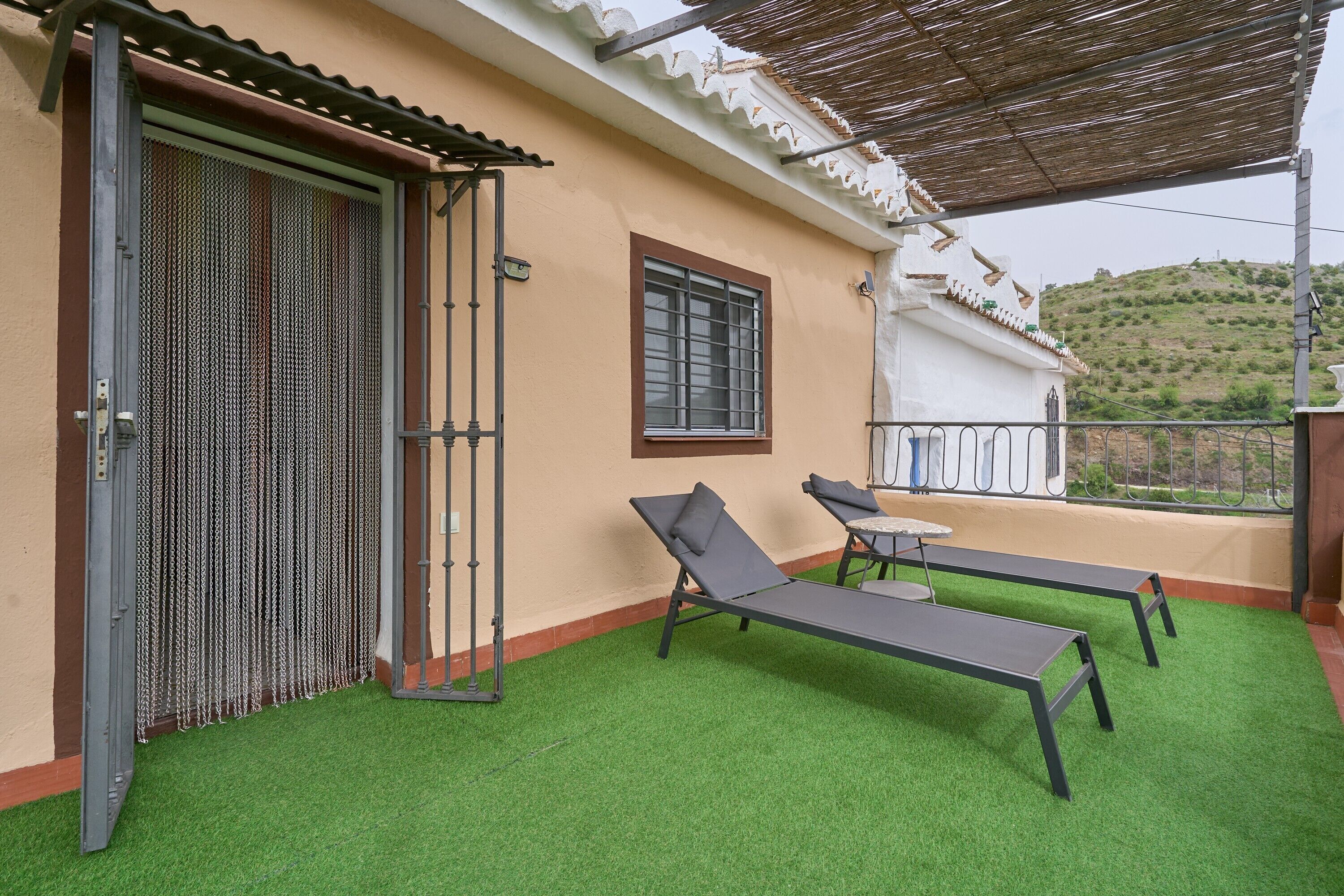 Een balkon met kunstgras, twee ligbedden, een tafeltje en een raam met een metalen traliewerk.