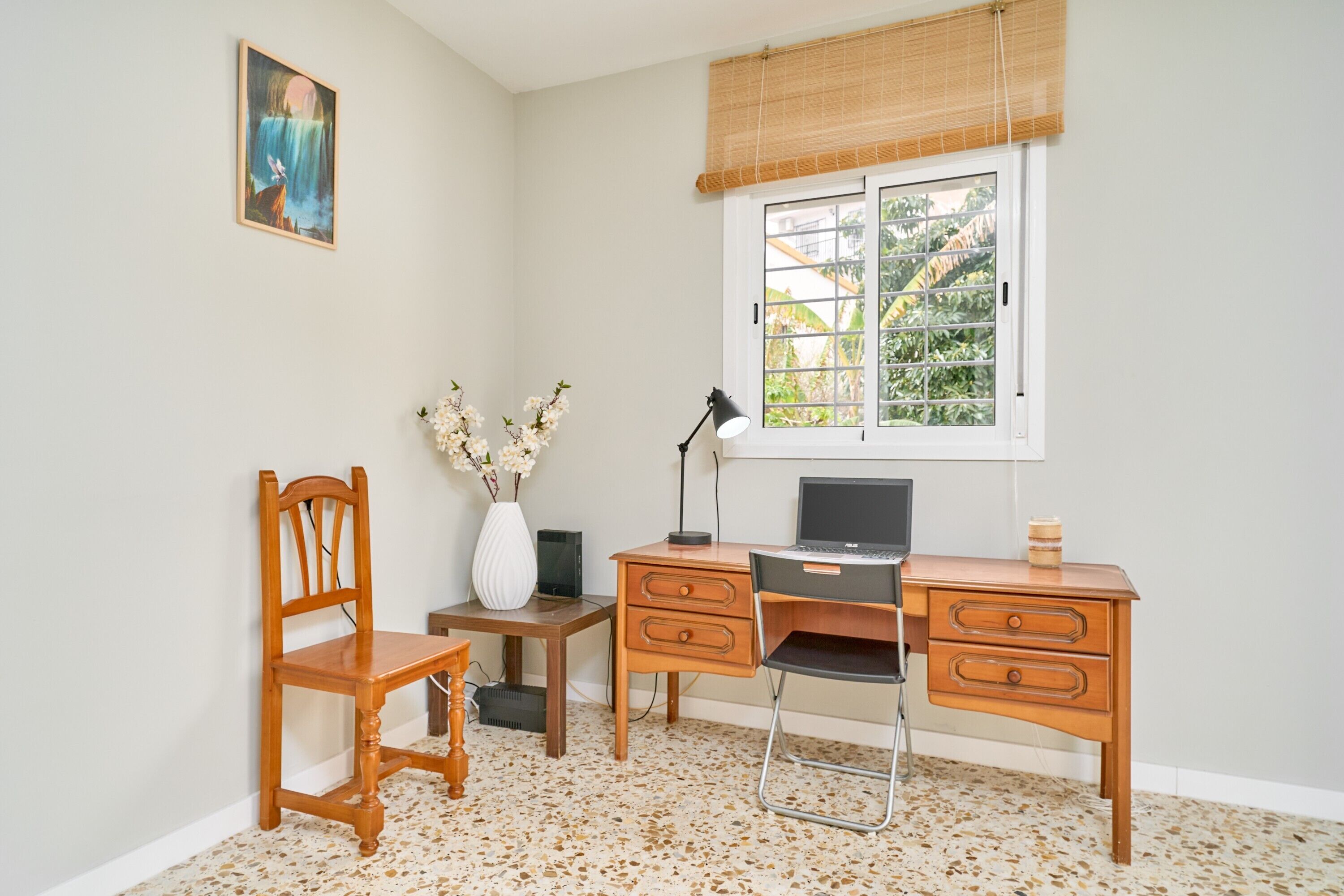 A room with a desk, chair, lamp, and a window with a view of greenery.