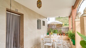 A covered patio with a dining table, chairs, potted plants, and a view of a hilly landscape.