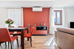 Een moderne woonkamer met een bank, een eettafel met stoelen, een keuken met kastjes, en een groot raam met uitzicht.