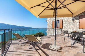 Een terras met een tafel en stoelen, een beige parasol, en een stenen gebouw met uitzicht op de zee.