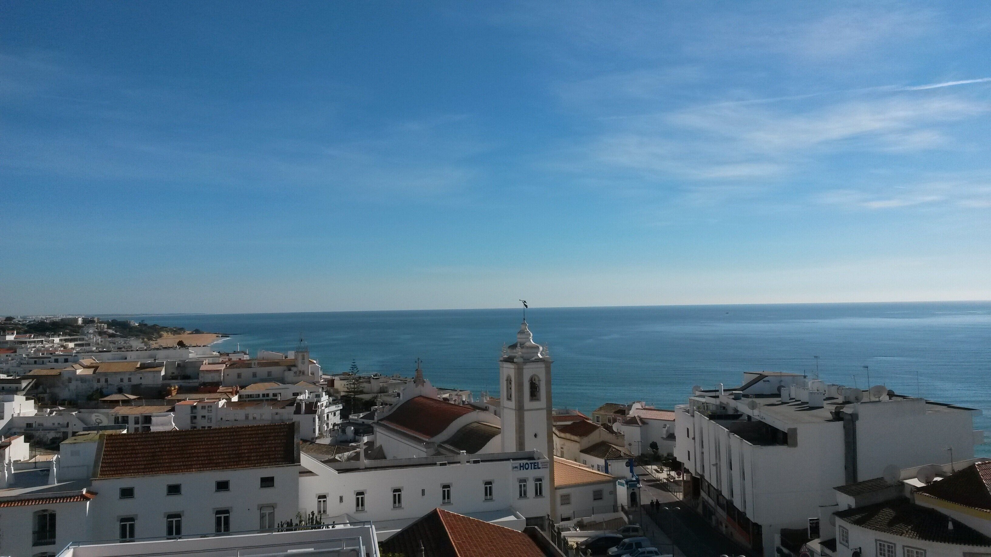 Un pueblo costero con una imponente torre de iglesia blanca, edificios blancos con tejados de tejas rojas y una vista despejada del mar.