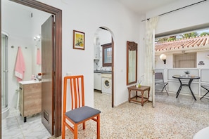 A room with a wooden chair, a small table with a lamp, a mirror, and a view of a kitchen area. - Apartment 'Calm Bungalow' with Shared Pool, Wi-Fi and Air Conditioning (Torrox)