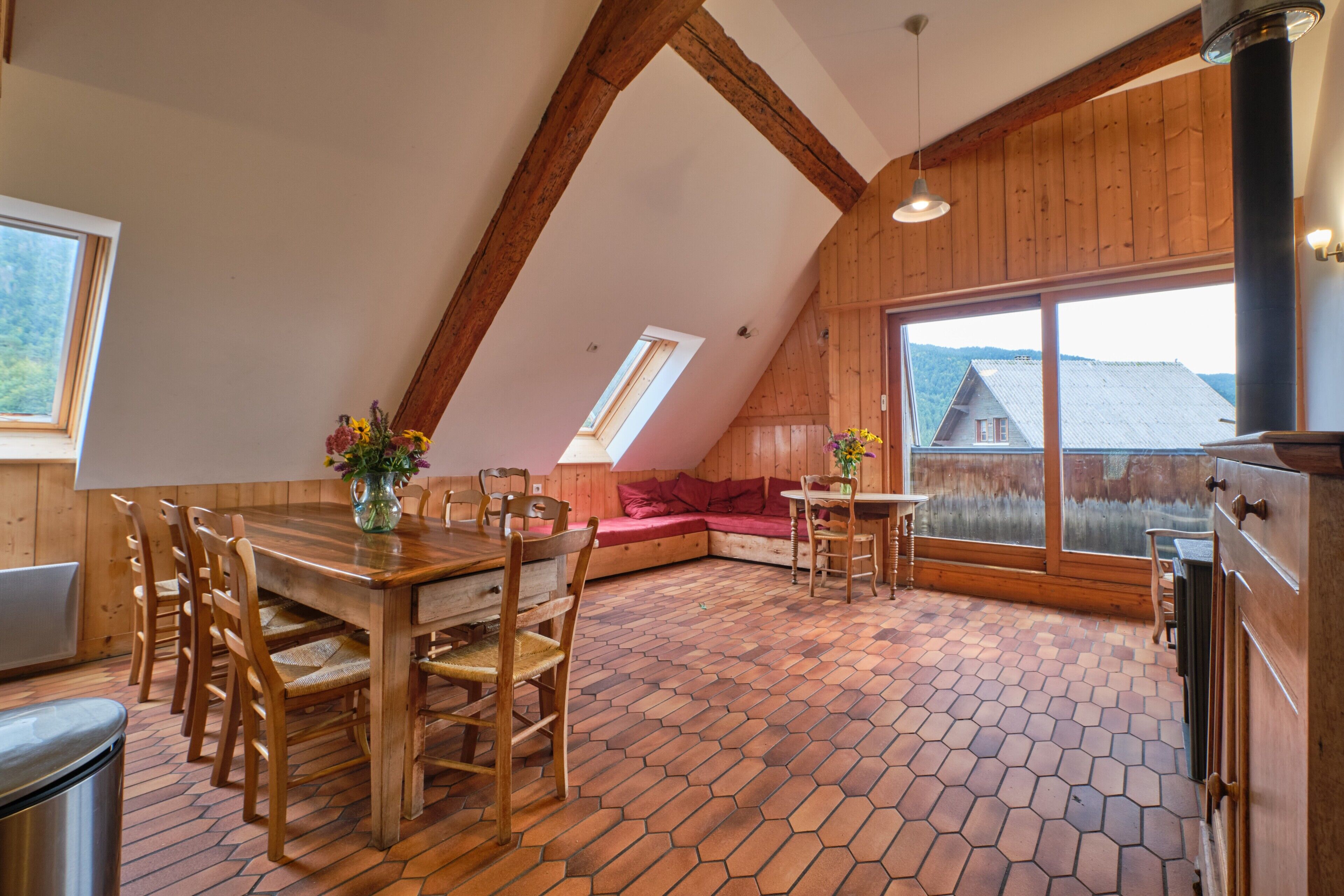 Een houten interieur met een eettafel, stoelen, een bank en een groot raam met uitzicht op een huis.