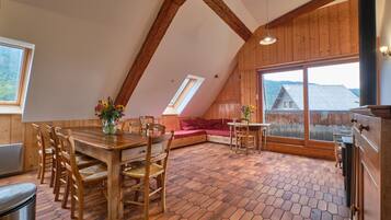 Een houten interieur met een eettafel, stoelen, een bank en een groot raam met uitzicht op een huis.