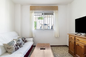 Ein Zimmer mit einem weißen Sofa, einem Holztisch, einem Fernseher auf einer Holzkommode und einem Fenster mit Blick auf Grünflächen.