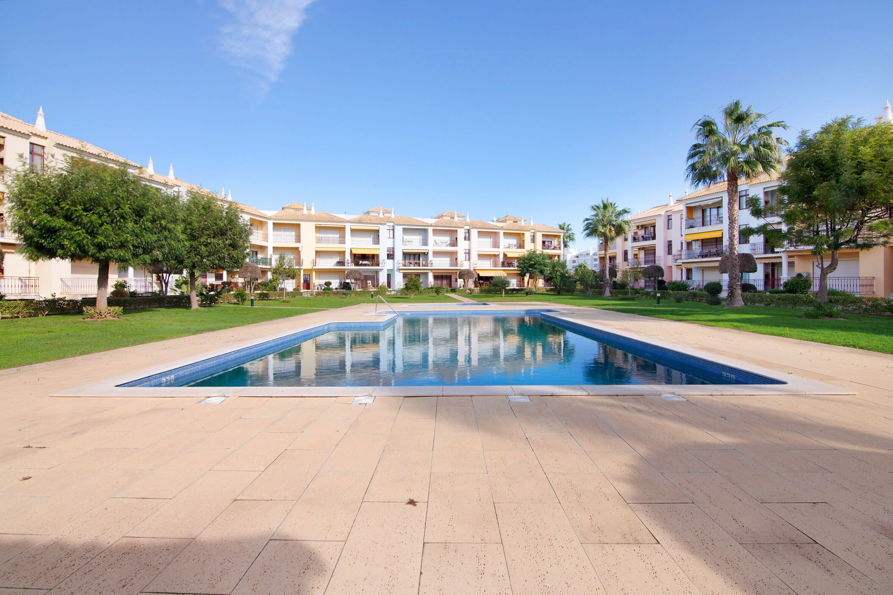 A swimming pool surrounded by a courtyard with buildings, trees, and a clear sky.