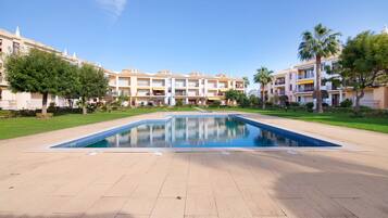 A swimming pool surrounded by a courtyard with buildings, trees, and a clear sky.