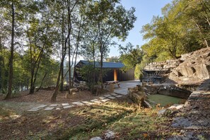 Una baita rustica con facciata in pietra, immersa in una zona boschiva con un sentiero in pietra che conduce a un portico di legno.