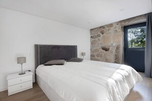 Une chambre avec un mur en pierre, un lit avec une literie blanche, une table de chevet avec une lampe et une fenêtre avec des rideaux.
