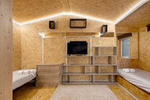 Una stanza compatta con TV integrata, pareti in legno, un letto bianco, una cassettiera in legno e una finestra.