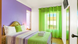 Une chambre avec un lit, une table de chevet, un téléviseur fixé au mur et une fenêtre avec des rideaux verts.