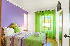 Une chambre avec un lit, une table de chevet, un téléviseur fixé au mur et une fenêtre avec des rideaux verts.