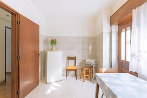 Une cuisine compacte comprenant un réfrigérateur, une chaise en bois, une petite table et une fenêtre avec des rideaux.