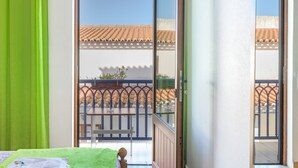 Une chambre avec un lit, un rideau vert, un balcon donnant sur un toit recouvert de tuiles et une rambarde en métal.