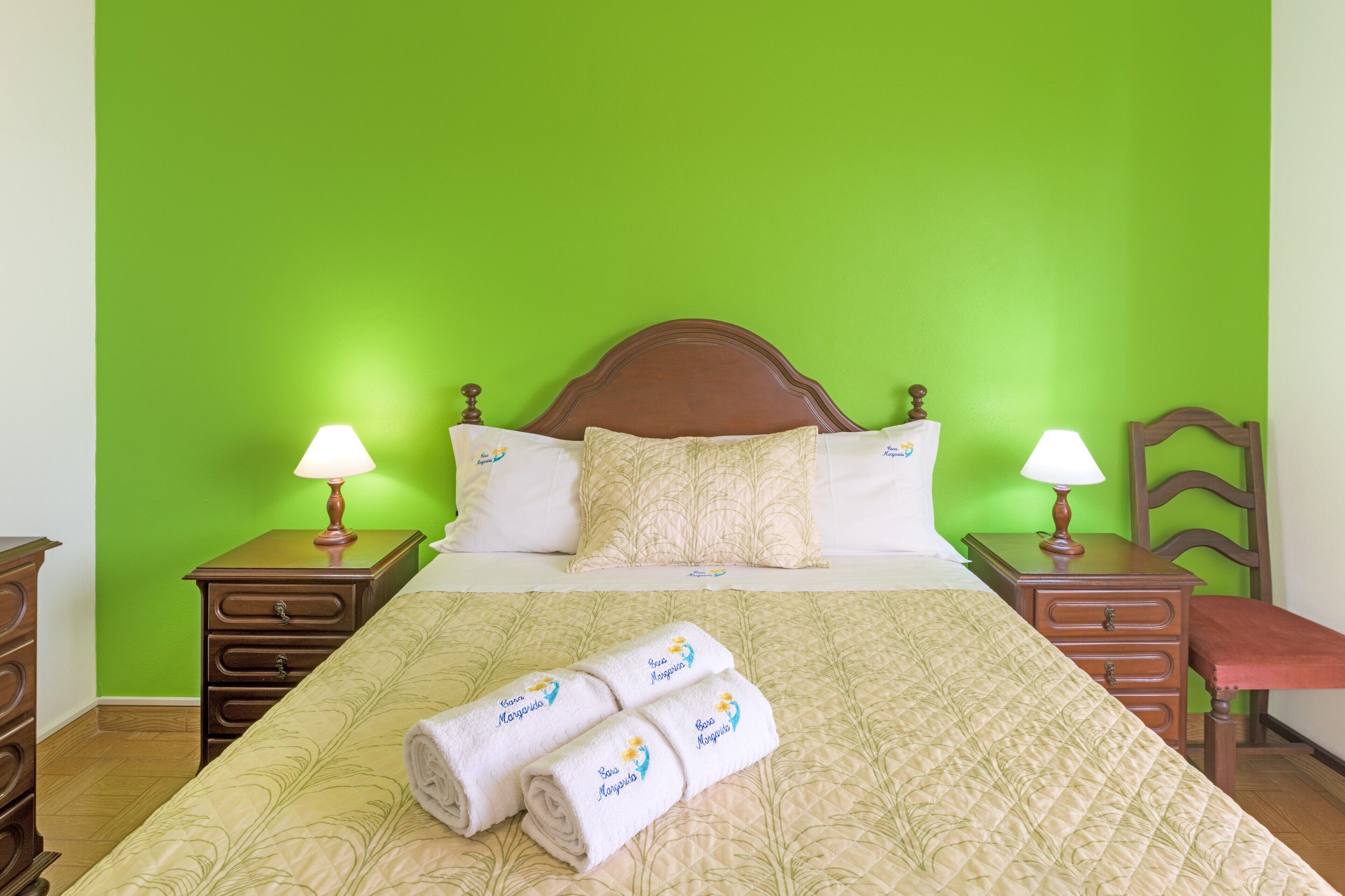 Une chambre d’hôtel avec un mur vert, une tête de lit en bois, un lit recouvert d’une courtepointe matelassée, deux tables de chevet avec des lampes et deux serviettes enroulées posées sur le lit.