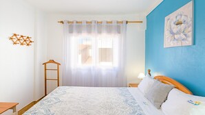 A bedroom with a bed, a wooden ladder, a window with sheer curtains, a blue wall, and a floral painting.