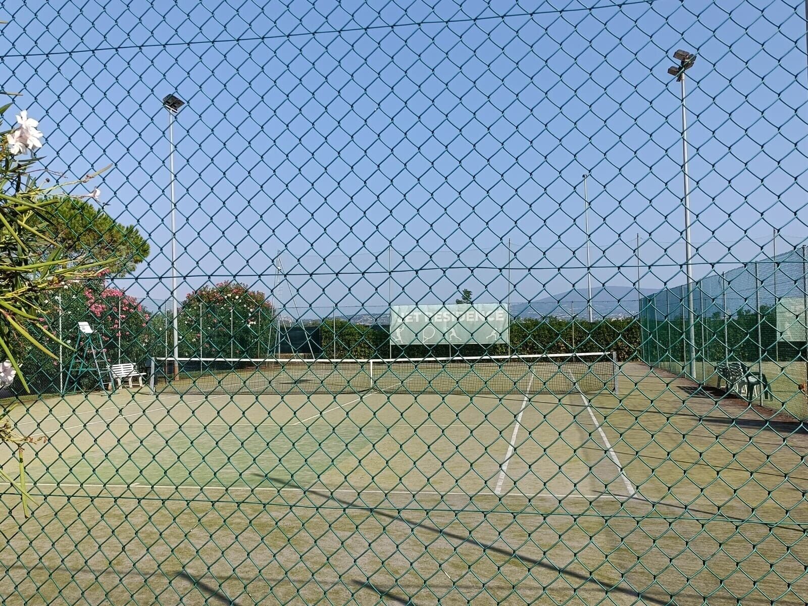 Een tennisbaan omringd door een hek van gaas, met een bankje en een bord in de verte.
