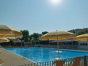 A swimming pool with multiple yellow striped umbrellas, a clear blue sky, and a building in the background.