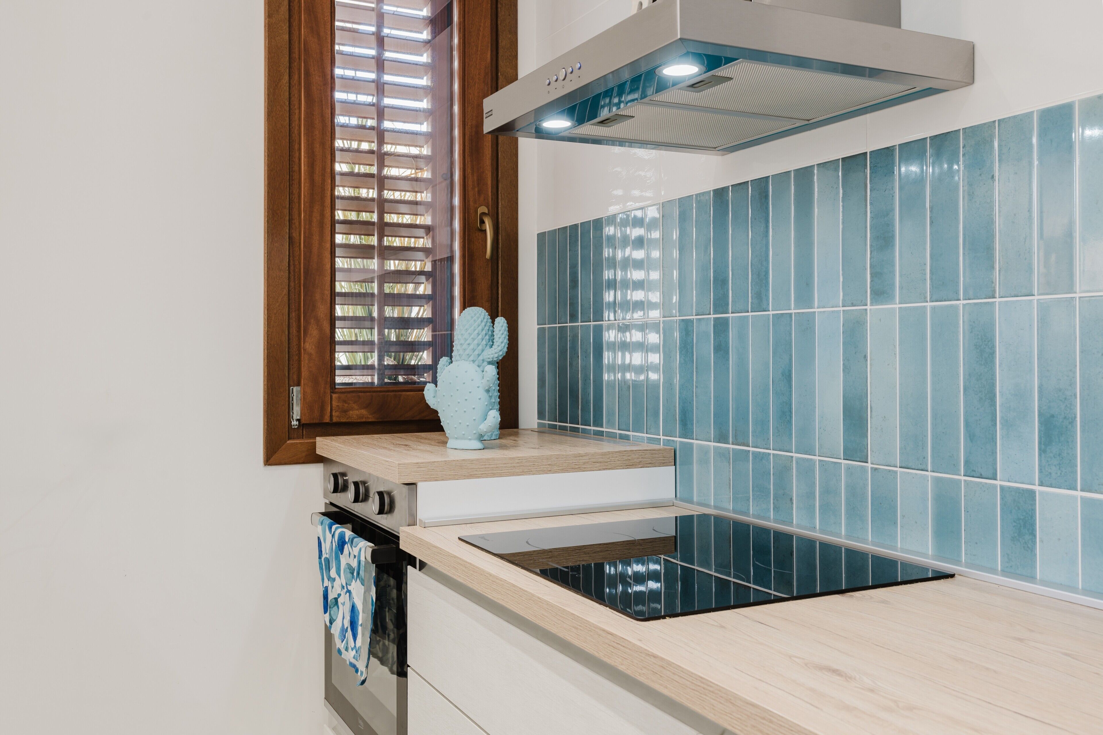 A modern kitchen with a built-in stove, a window with blinds, and blue-tiled walls.