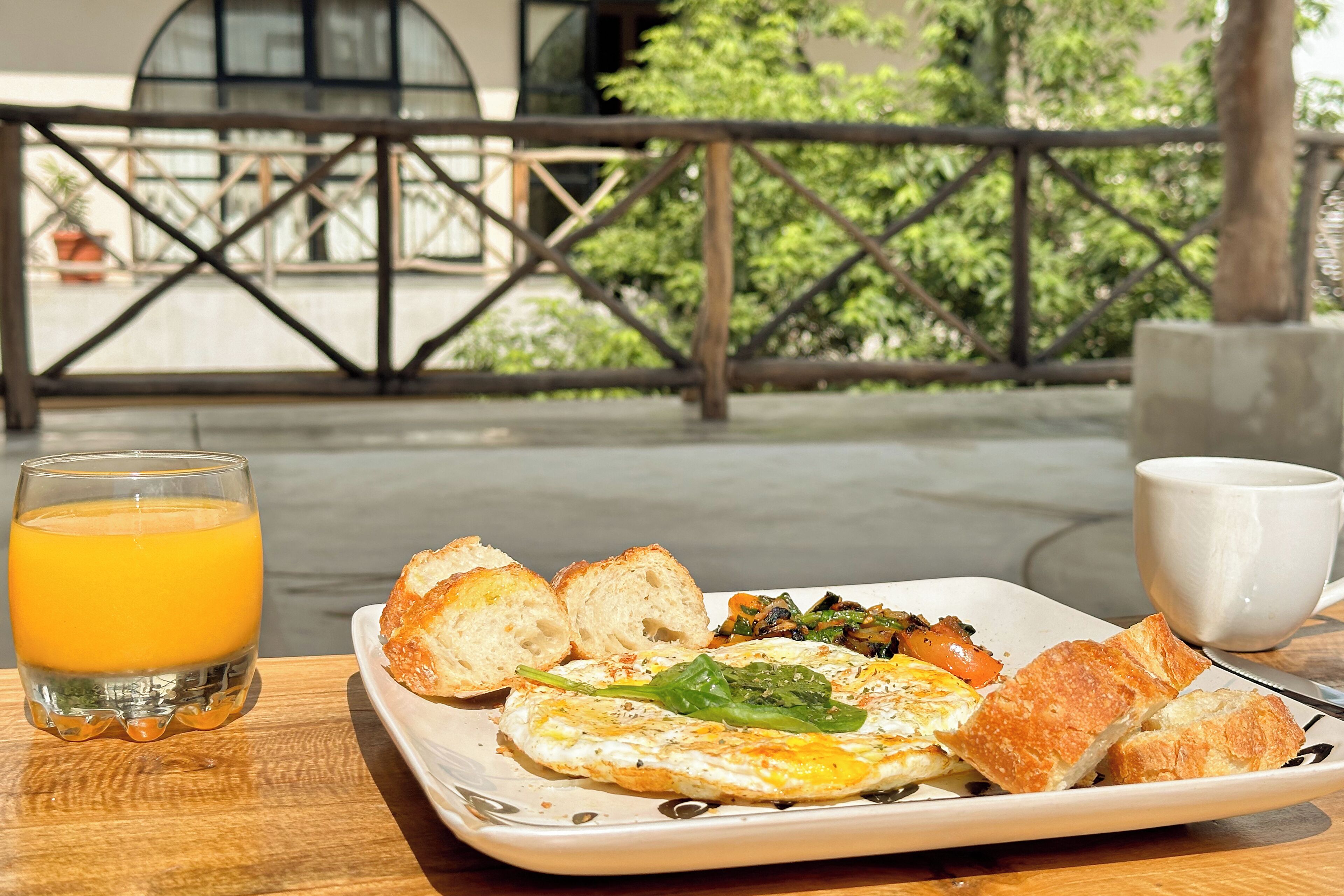 Bandeja de desayuno con huevos, tostadas, vegetales, un vaso de jugo de naranja y una taza blanca sobre una mesa de madera.