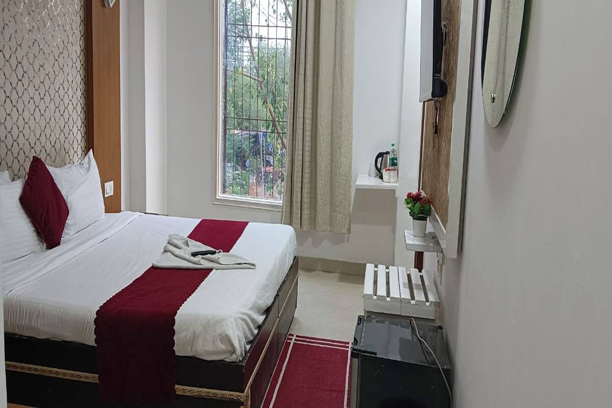 A hotel room with a bed, a window with a view of greenery, a small shelf with a kettle, and a radiator.