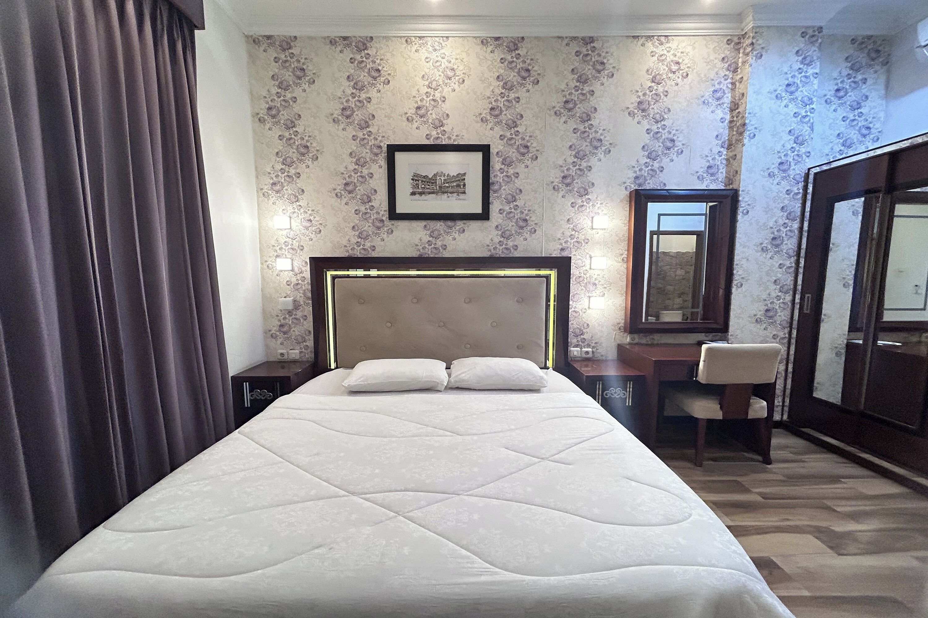 A hotel room with a large bed, a desk with a chair, a mirror, and a framed picture on the wall.