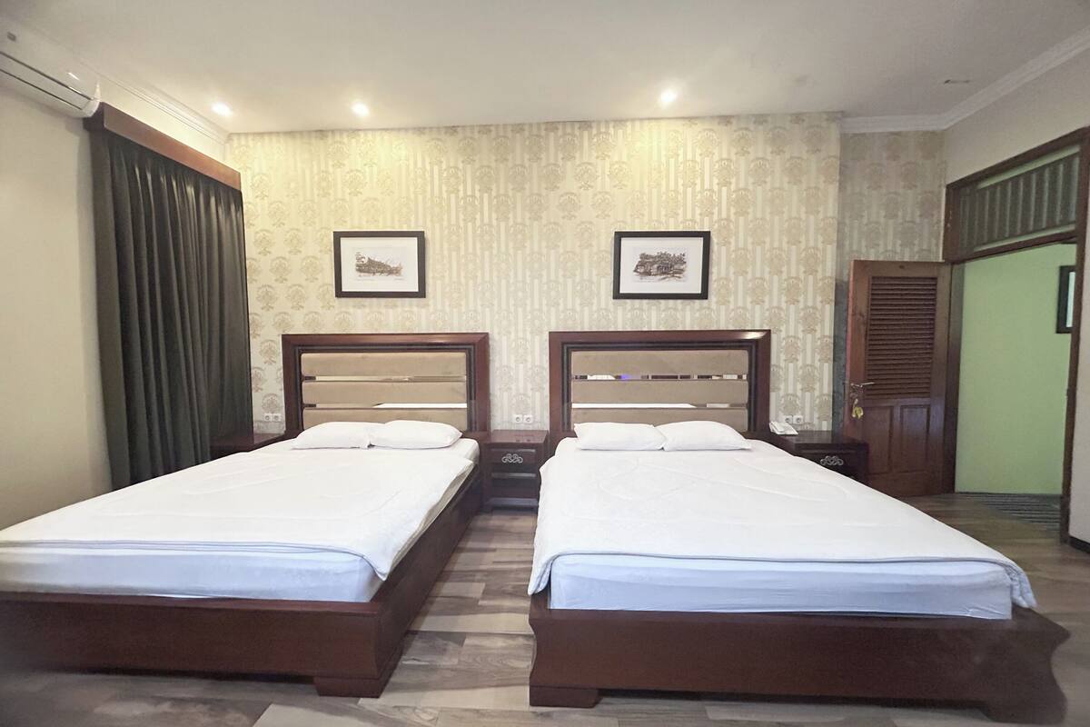 A hotel room with two single beds, a wooden headboard, a small bedside table, a nightstand, a framed picture on the wall, and a door with a green frame.