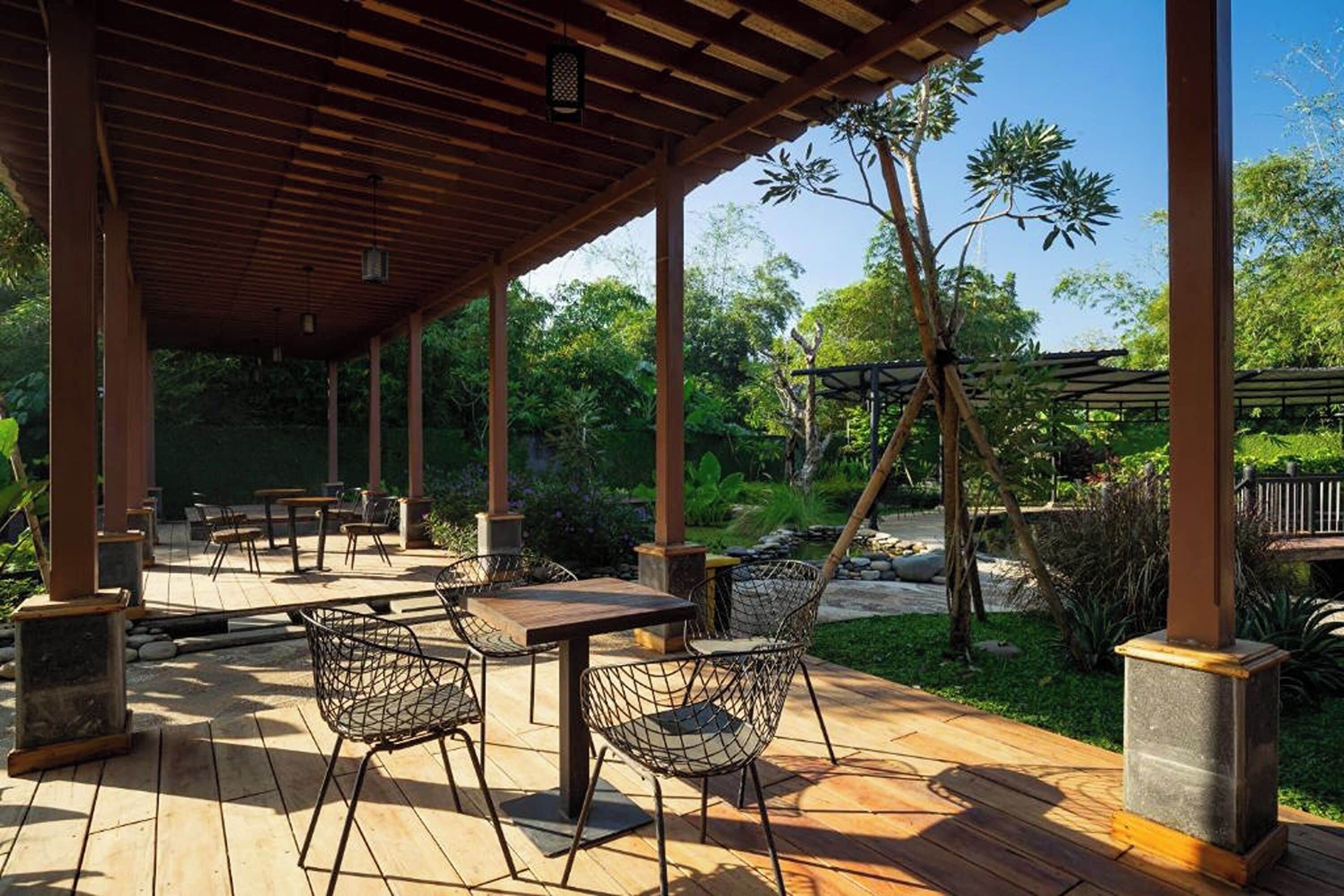 An outdoor patio area with wooden tables and chairs, surrounded by greenery and a pergola structure.
