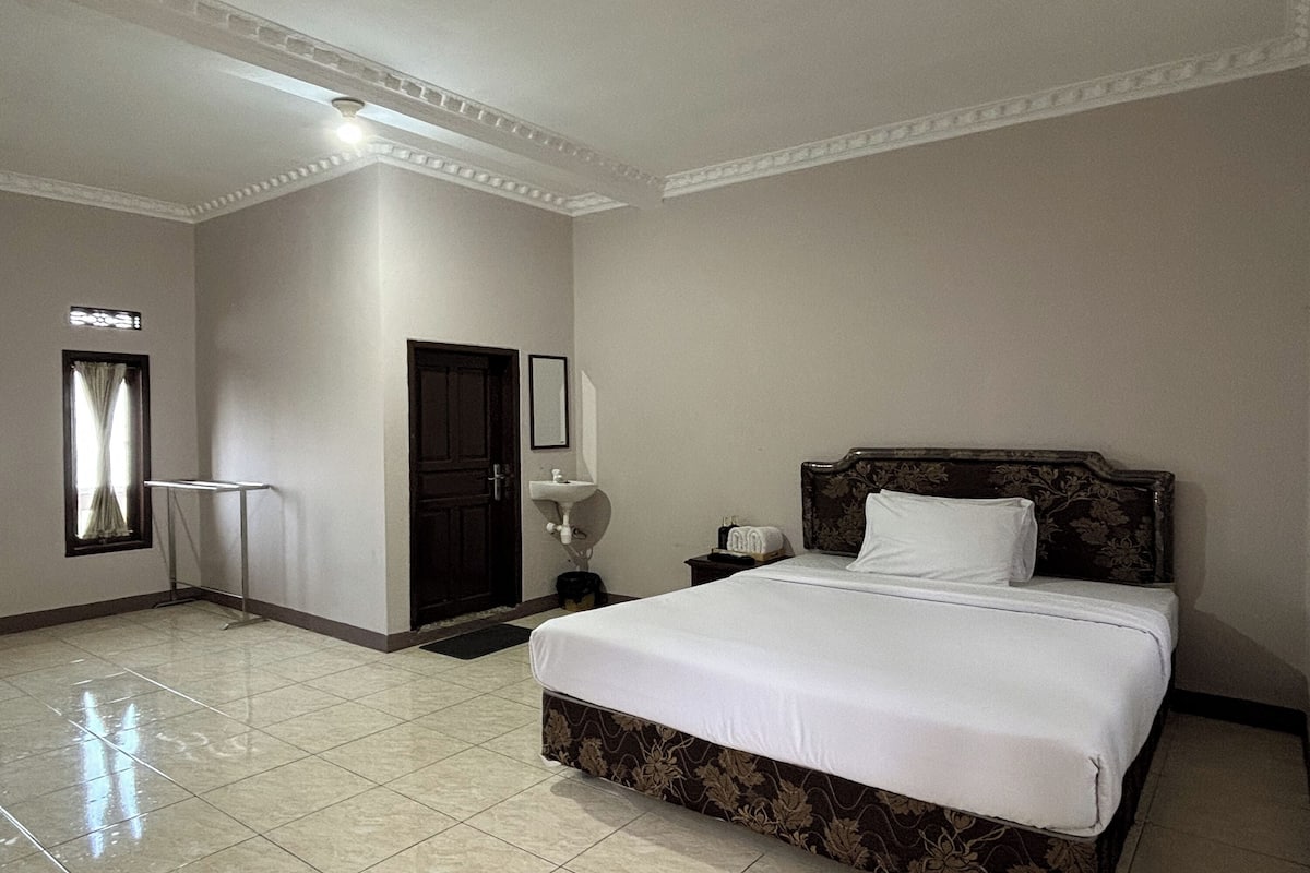 Quarto de hotel com uma cama grande, uma mesa de cabeceira, cortinas numa janela e uma porta.