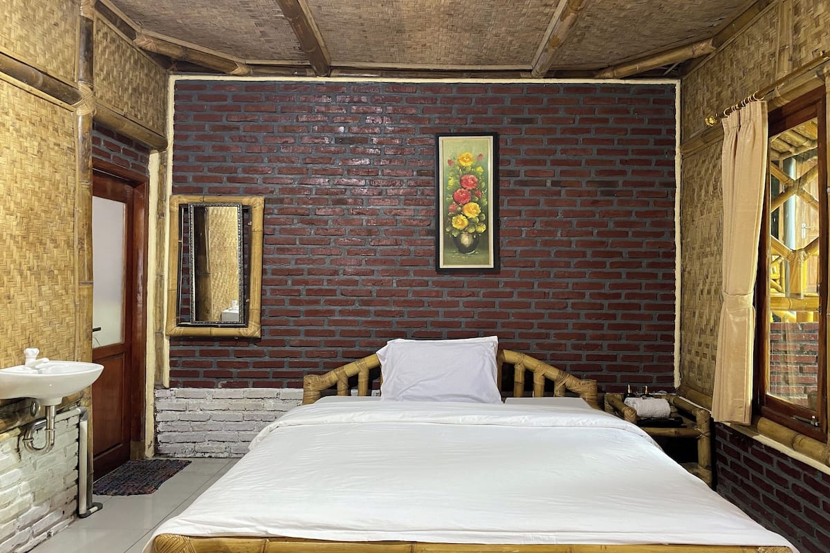 Quarto com parede de tijolos, cama com roupa de cama branca, cabeceira de madeira, mesa de cabeceira de madeira, pia branca e um quadro floral emoldurado.