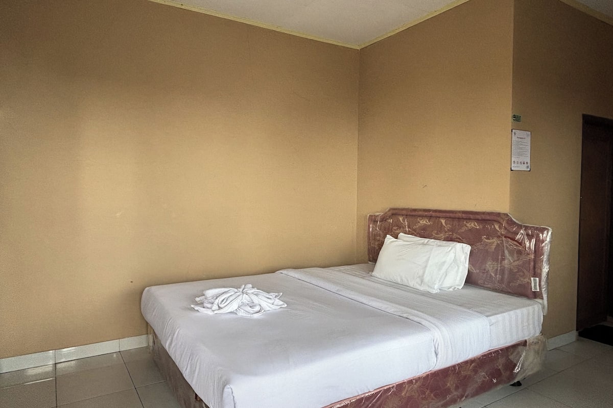 Uma cama de solteiro com colchão e almofadas brancos, uma toalha branca dobrada e um encosto com capa de plástico.