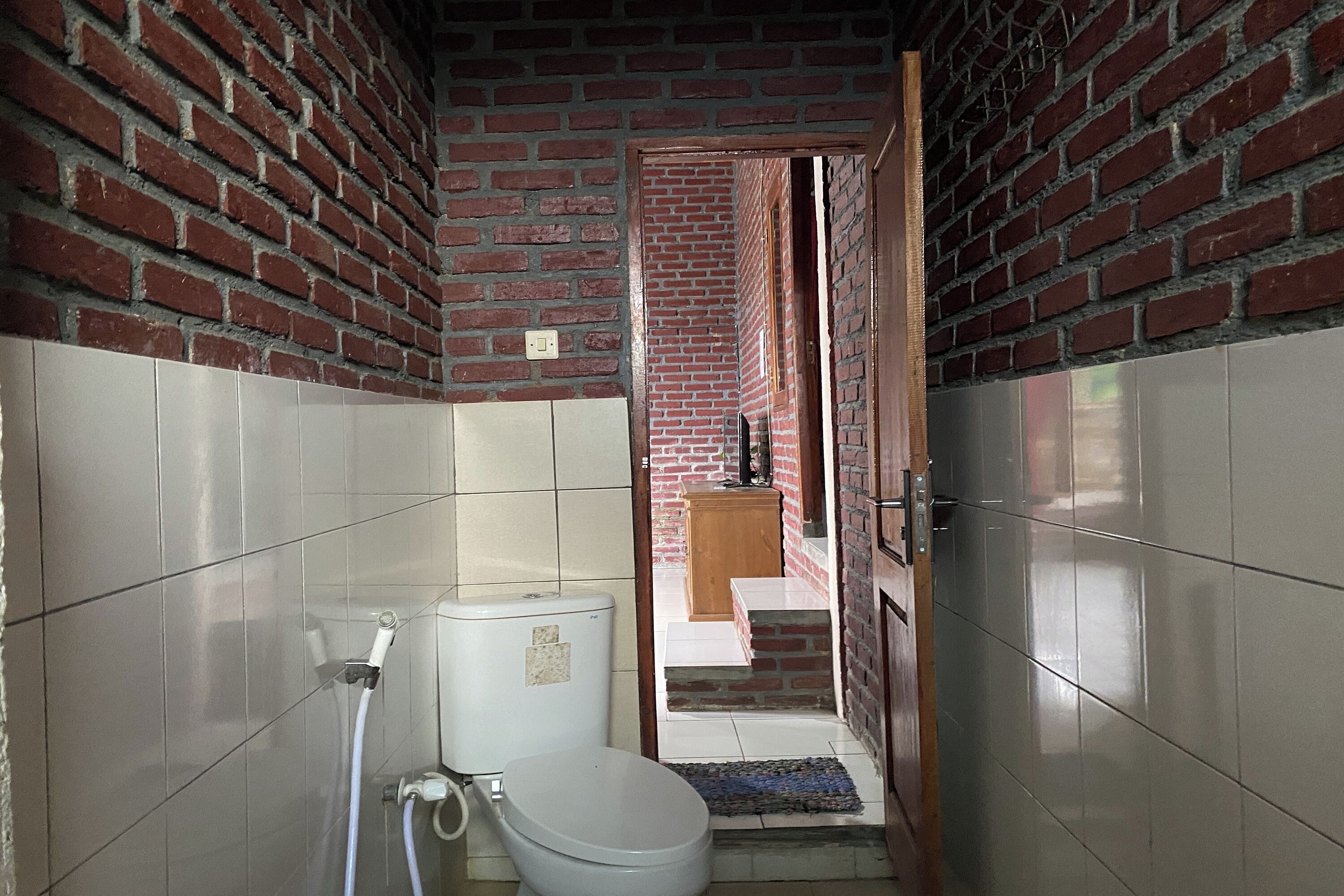 Bany amb vàter, dutxa i rentapans. Una paret revestida de rajoles i una paret de maó. Una porta condueix a una altra habitació.