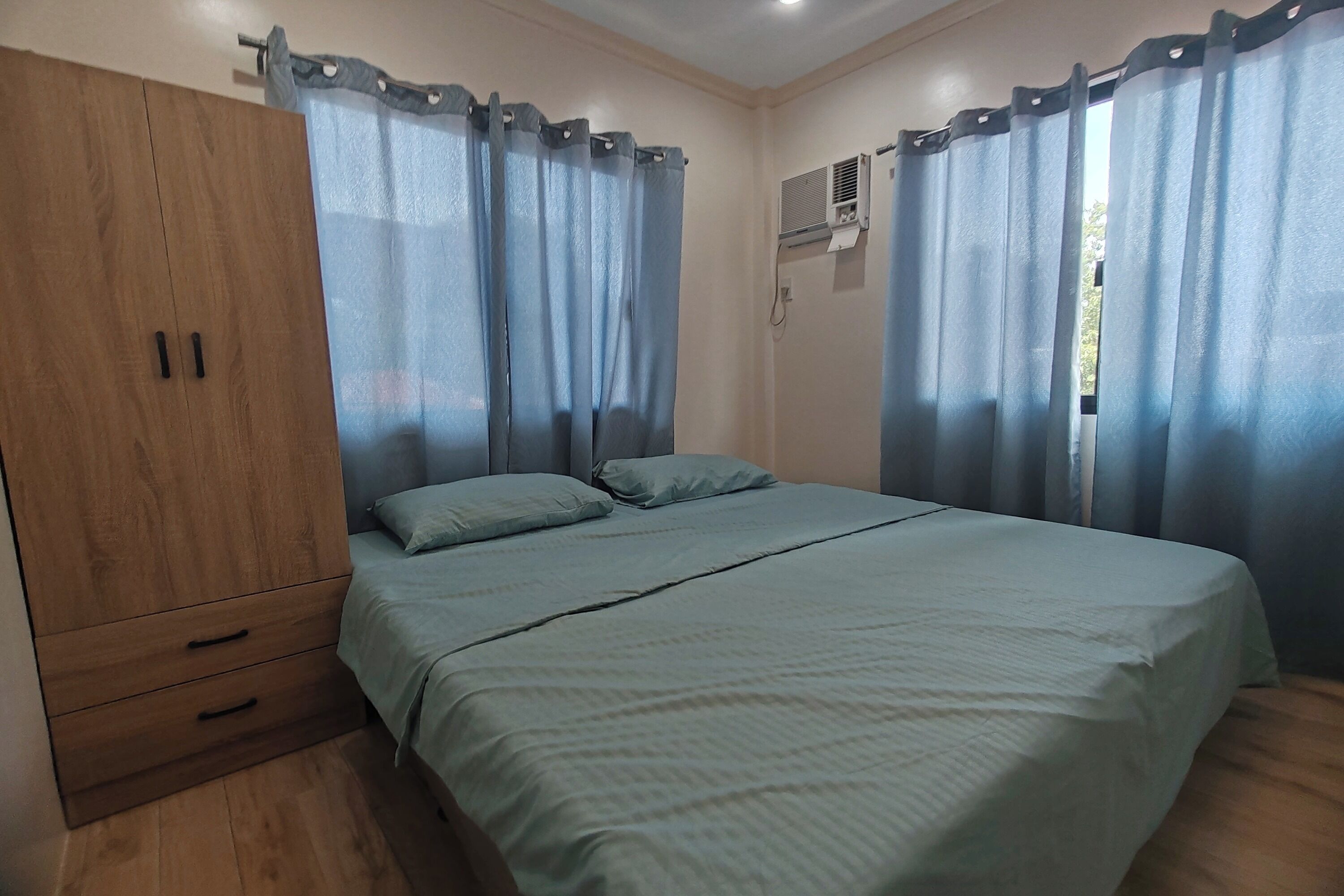 A bedroom with a wooden wardrobe, a bed with a green bedspread, and blue curtains.