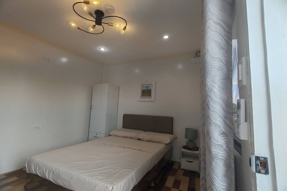 A bedroom with a bed, a bedside table with a lamp, a ceiling light fixture, and a door.